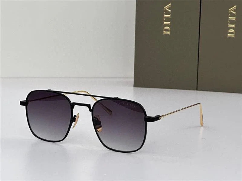 Black and gold square sunglasses with gradient lenses on a white surface, luxury eyewear box behind.