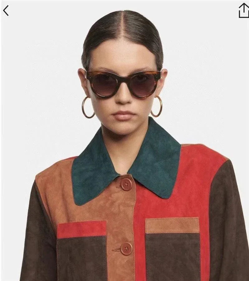 Woman wearing round oversized sunglasses, gold hoop earrings, and a colorblock suede jacket