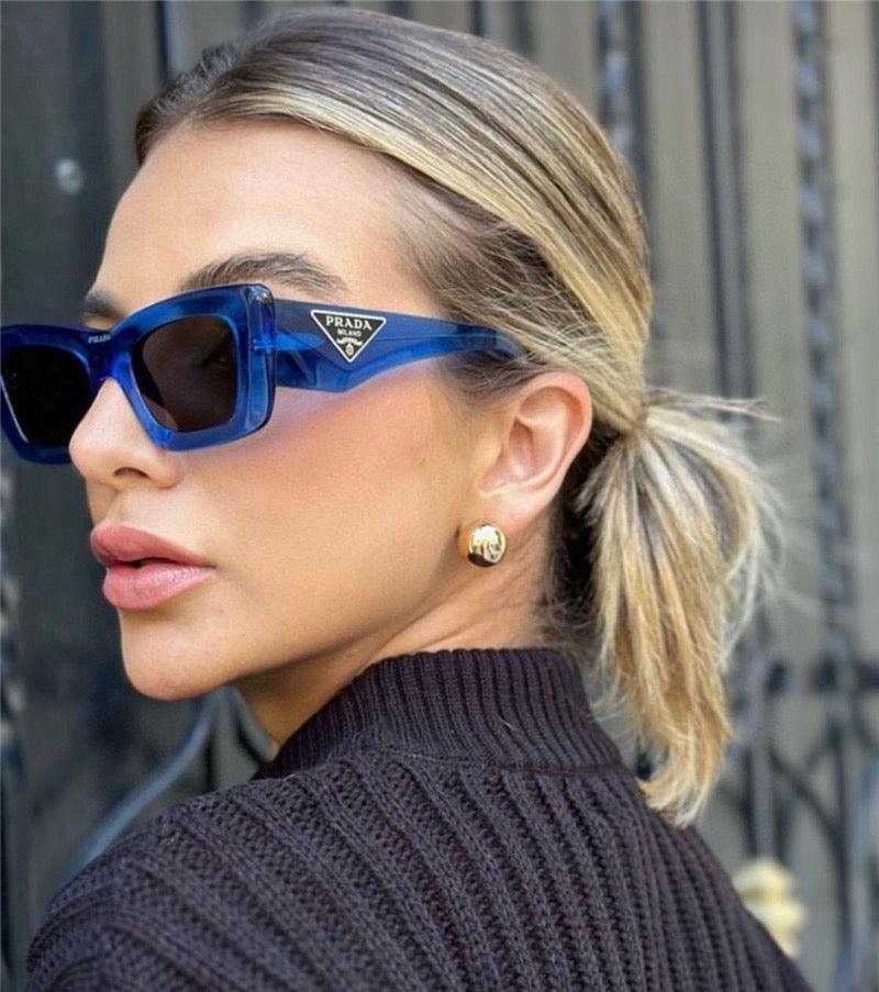 Woman wearing blue square Prada sunglasses and gold stud earrings outdoors