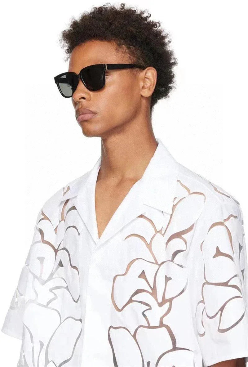 Man wearing black rectangular sunglasses and a white floral cut-out shirt, isolated background.